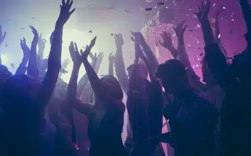 Menschen tanzen mit erhobenen Händen unter bunten Lichtern und Konfetti, während ein DJ aus Berlin auf einer lebhaften Party oder in einem Nachtclub für Stimmung sorgt.
