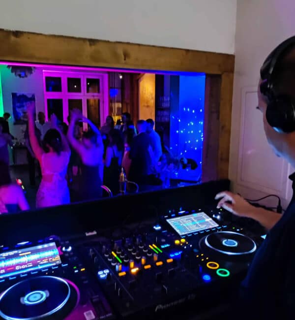 Der Hochzeits-DJ Oliver Scheibner heizt eine lebhafte Party in Brandenburg an und legt Musik auf, während die Gäste unter bunten Lichtern im Hintergrund tanzen.