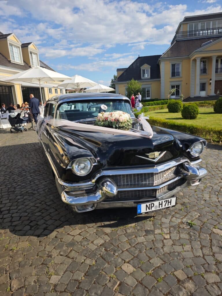 Vor eleganten Gebäuden steht ein schwarzer Oldtimer, der mit Blumen geschmückt ist, auf einer gepflasterten Einfahrt.