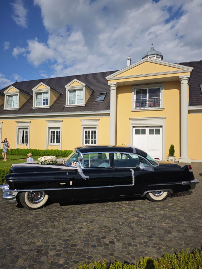 Ein klassisches schwarzes Auto steht auf einer gepflasterten Einfahrt vor einem großen gelben Haus mit weißen Säulen.