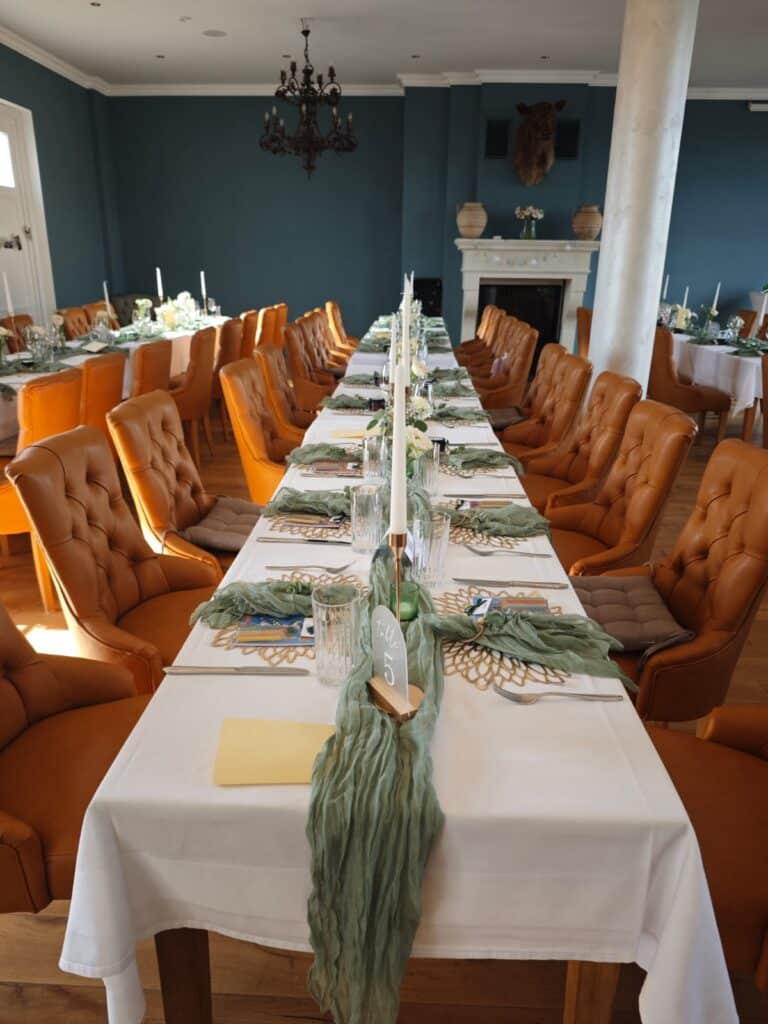 Ein langer Tisch, gedeckt für ein festliches Abendessen, mit orangefarbenen Stühlen und grüner Dekoration in einem Raum mit blauen Wänden.