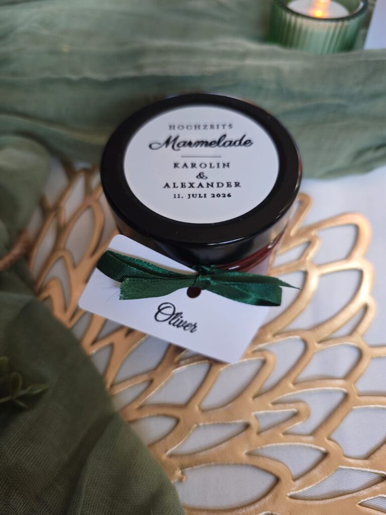 Ein Marmeladenglas zur Hochzeit mit der Aufschrift „Karolin & Alexander, 11. Juli 2026“ und einem Anhänger mit der Aufschrift „Oliver“.