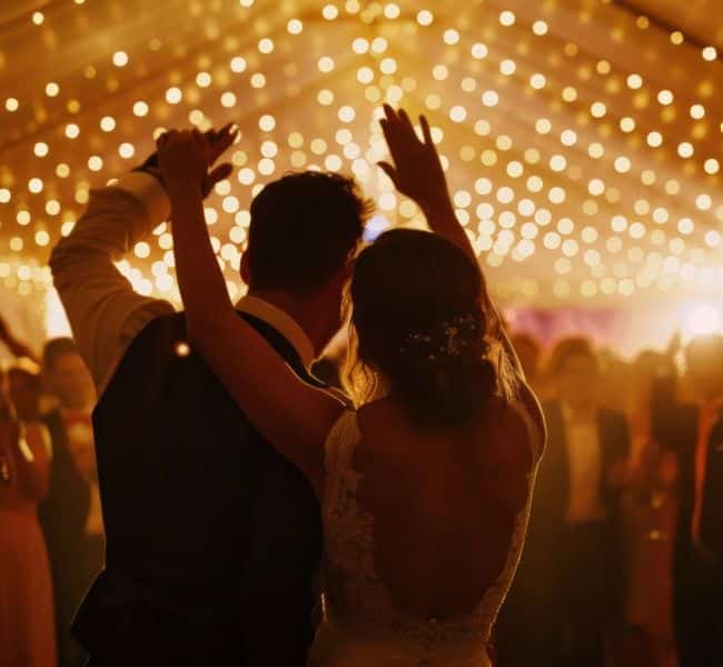 Ein Paar tanzt bei einer Hochzeit unter Lichterketten und reckt die Hände in die Höhe, während die Gäste zusehen und mitfeiern.