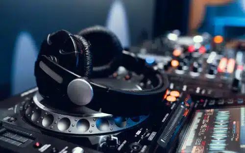 Ein schwarzer Kopfhörer liegt auf einem DJ-Mischpult, im Hintergrund sind bunte Lichter und Bedienelemente für die Audiosteuerung zu sehen.