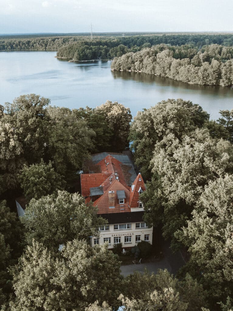 Luftaufnahme eines Gebäudes mit rotem Dach, umgeben von Bäumen in der Nähe eines großen Sees unter bewölktem Himmel.