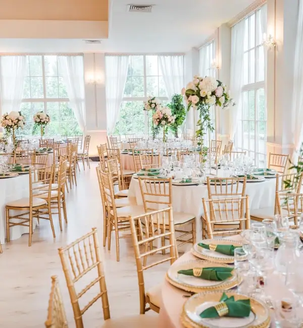 Ein eleganter Bankettsaal mit runden Tischen, goldenen Stühlen, grünen Servietten und hohen Blumengestecken.