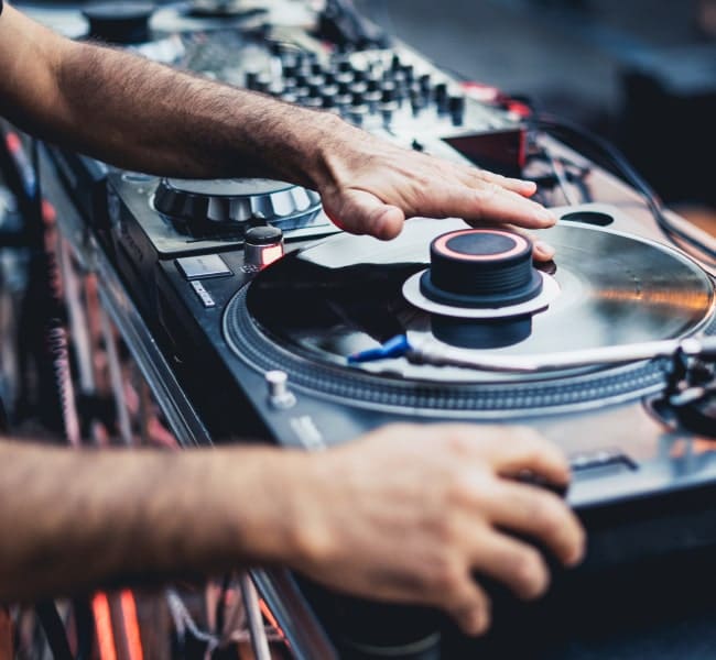 Die Hände eines DJs, der einen Plattenspieler einstellt; im Hintergrund ist die Mischanlage zu sehen.