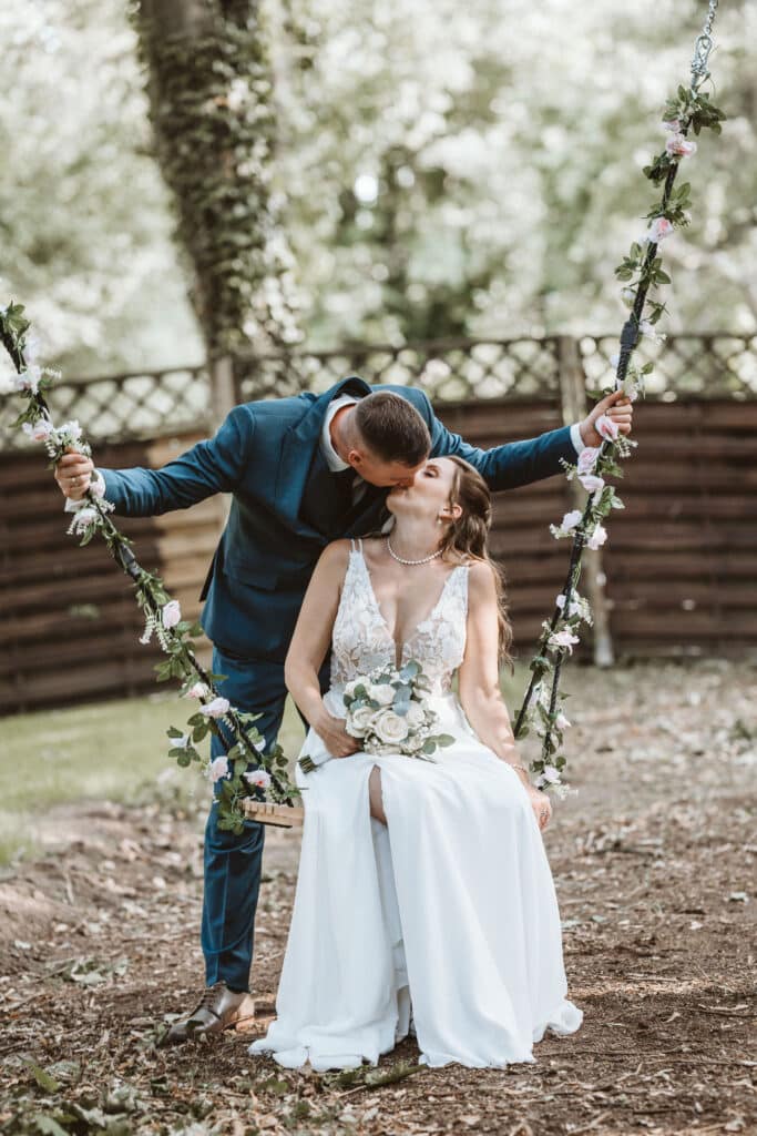 Der Bräutigam küsst die Braut, die auf einer mit Blumen geschmückten Schaukel sitzt und einen Blumenstrauß in der Hand hält – im Freien.