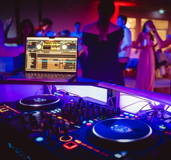 DJ-Ausrüstung und Laptop mit bunten Lichtern in einem Party-Lokal, im Hintergrund tanzen Menschen.
